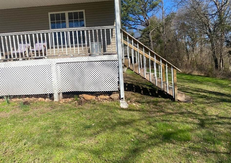 Porch with white railing and stairs beside a house, set in a grassy yard with trees.