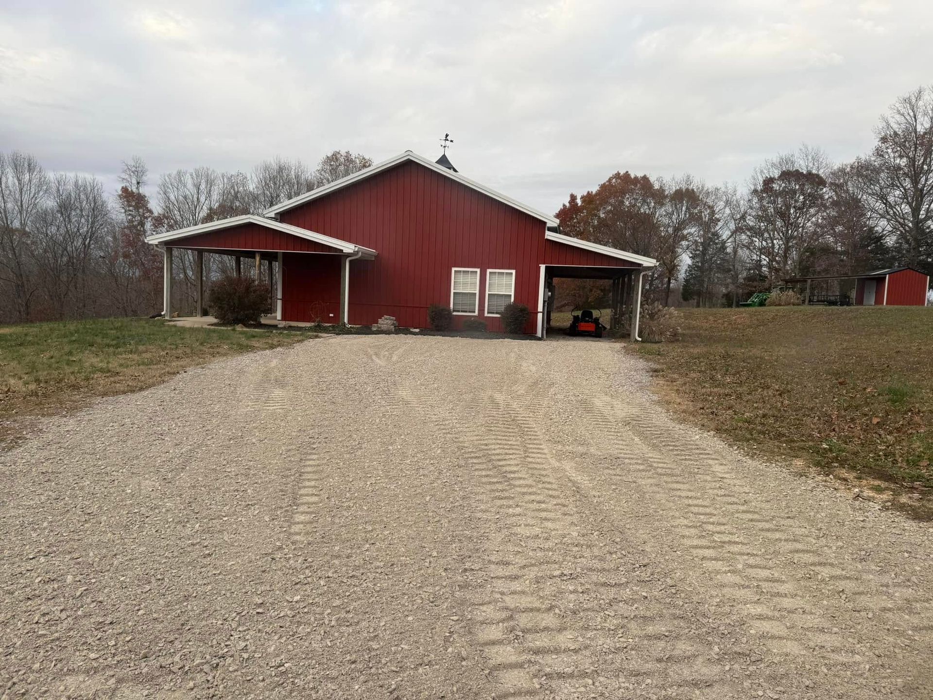 Red barn with covered side porches at the end of a gravel driveway, surrounded by bare trees.