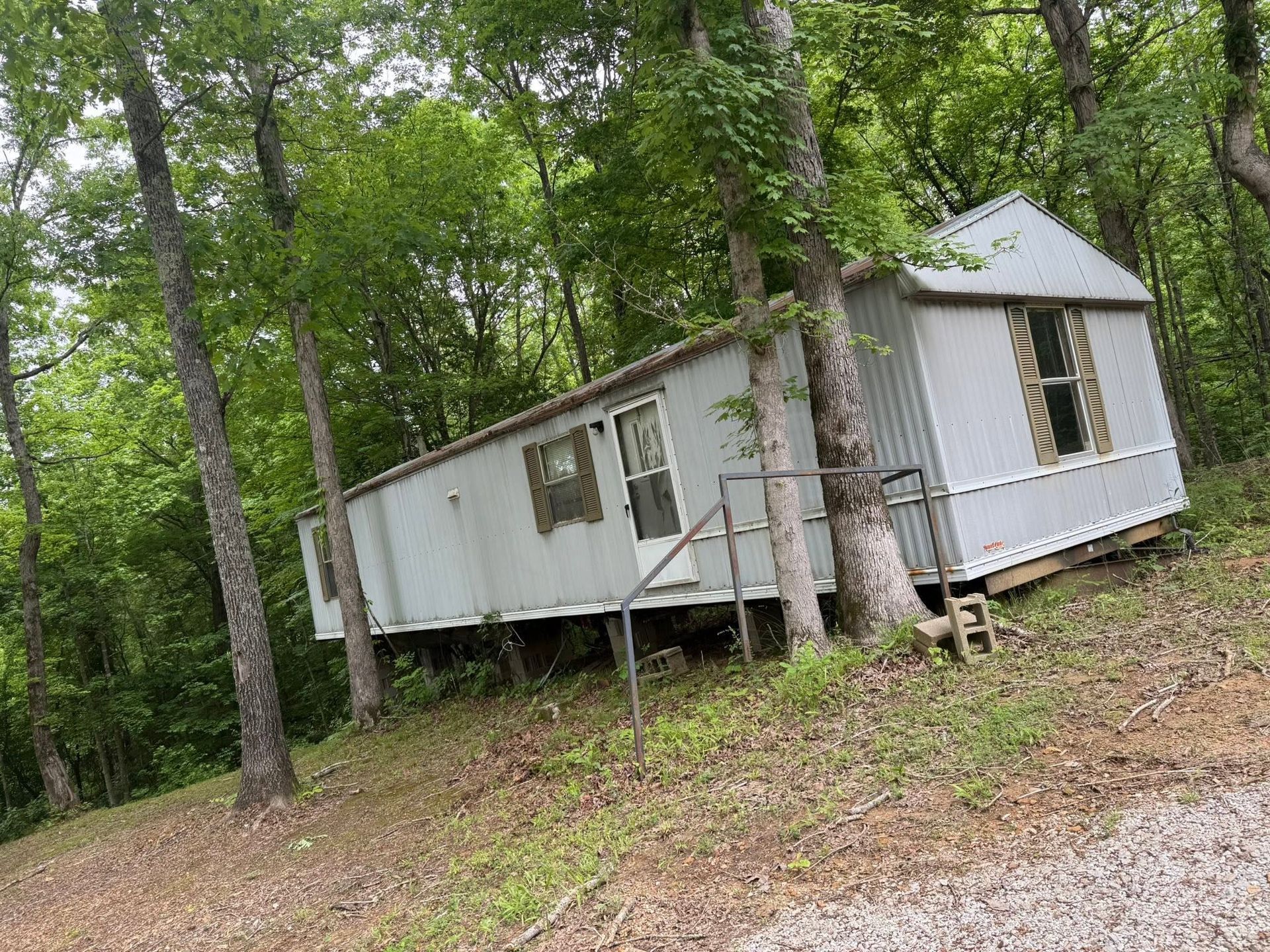 White cabin on stilts in a wooded area, with trees and a gravel clearing nearby.