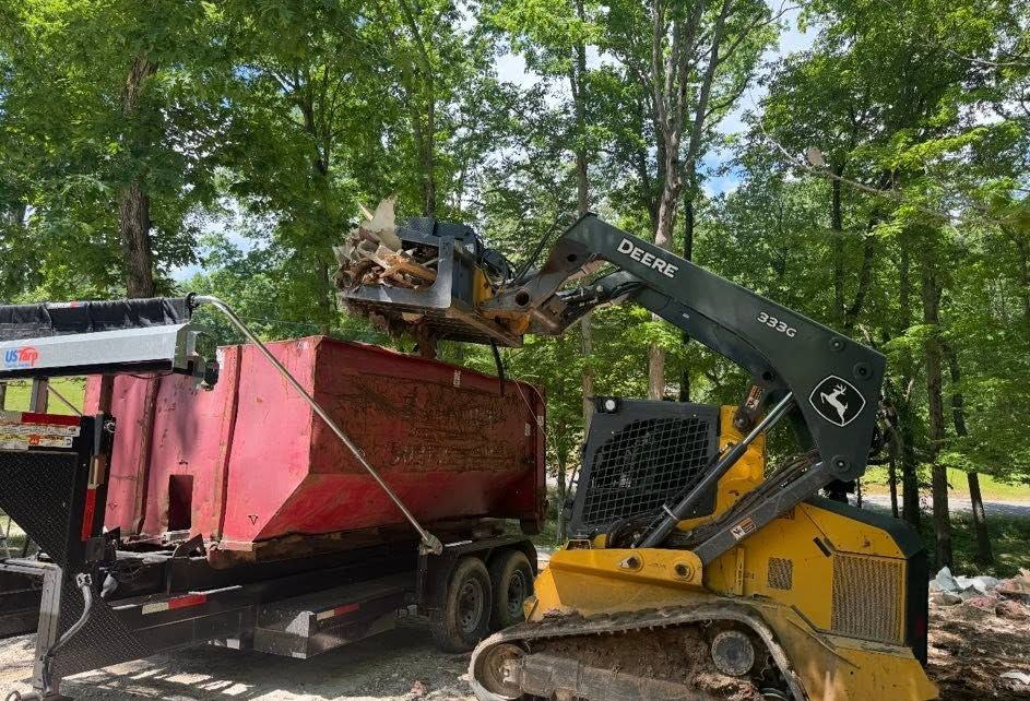 Yellow excavator loading debris into a red dump trailer in a wooded area