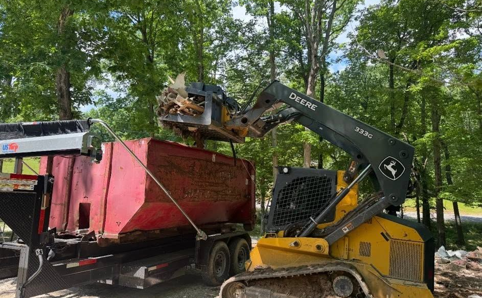Yellow skid steer loading debris into a red dumpster on a trailer in a wooded area.