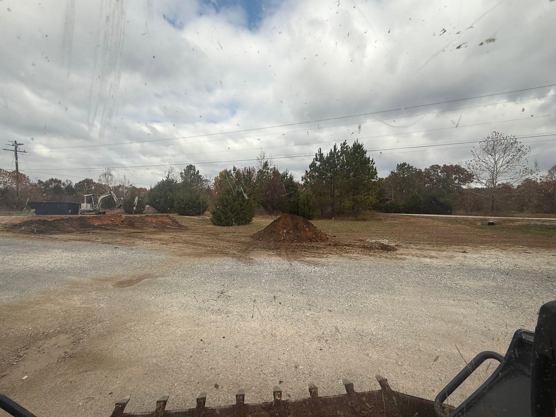 Gravel lot with dirt mounds and trees under a cloudy sky, viewed from a vehicle.