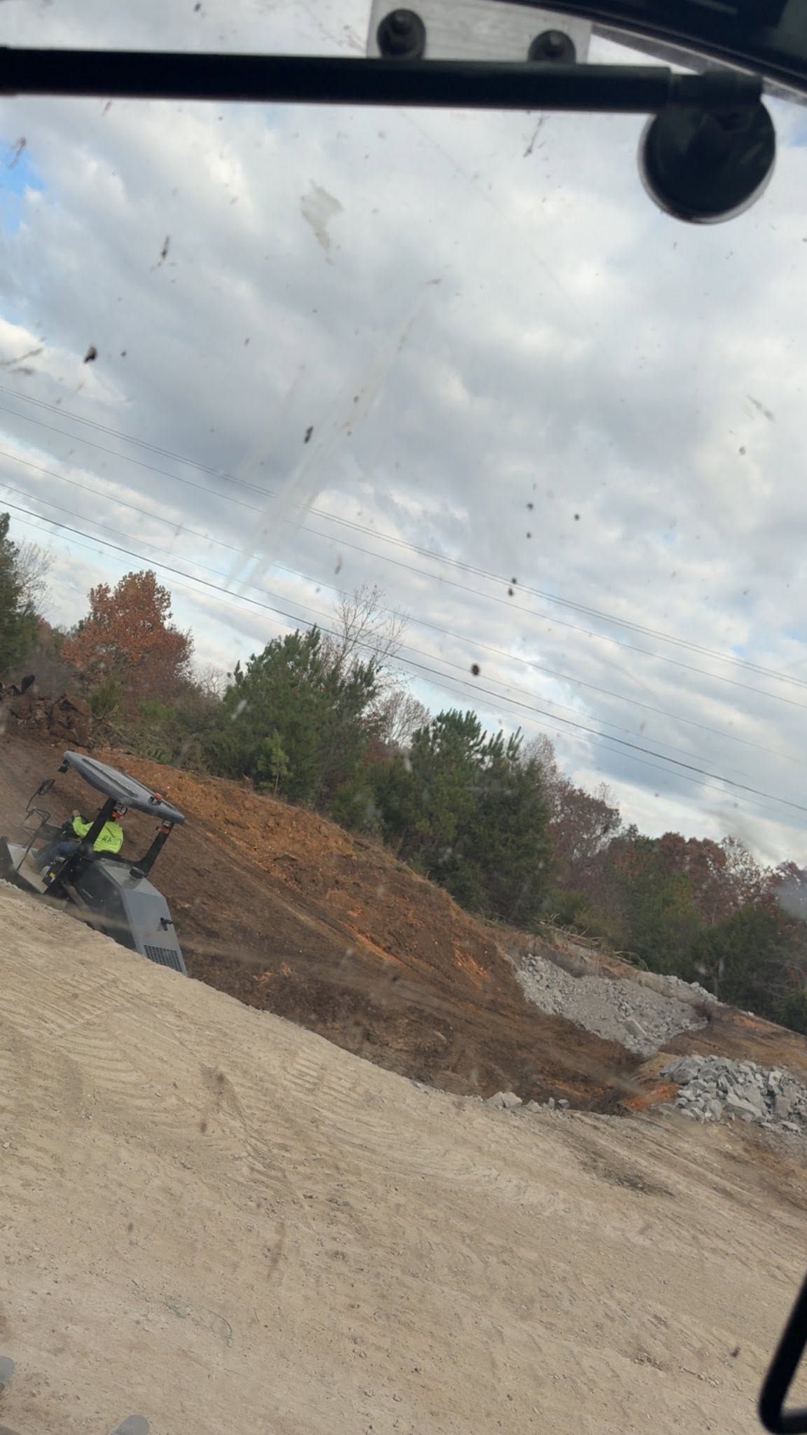 Tilted roadside view of a gravel pile, excavator, and cloudy sky seen through a vehicle windshield