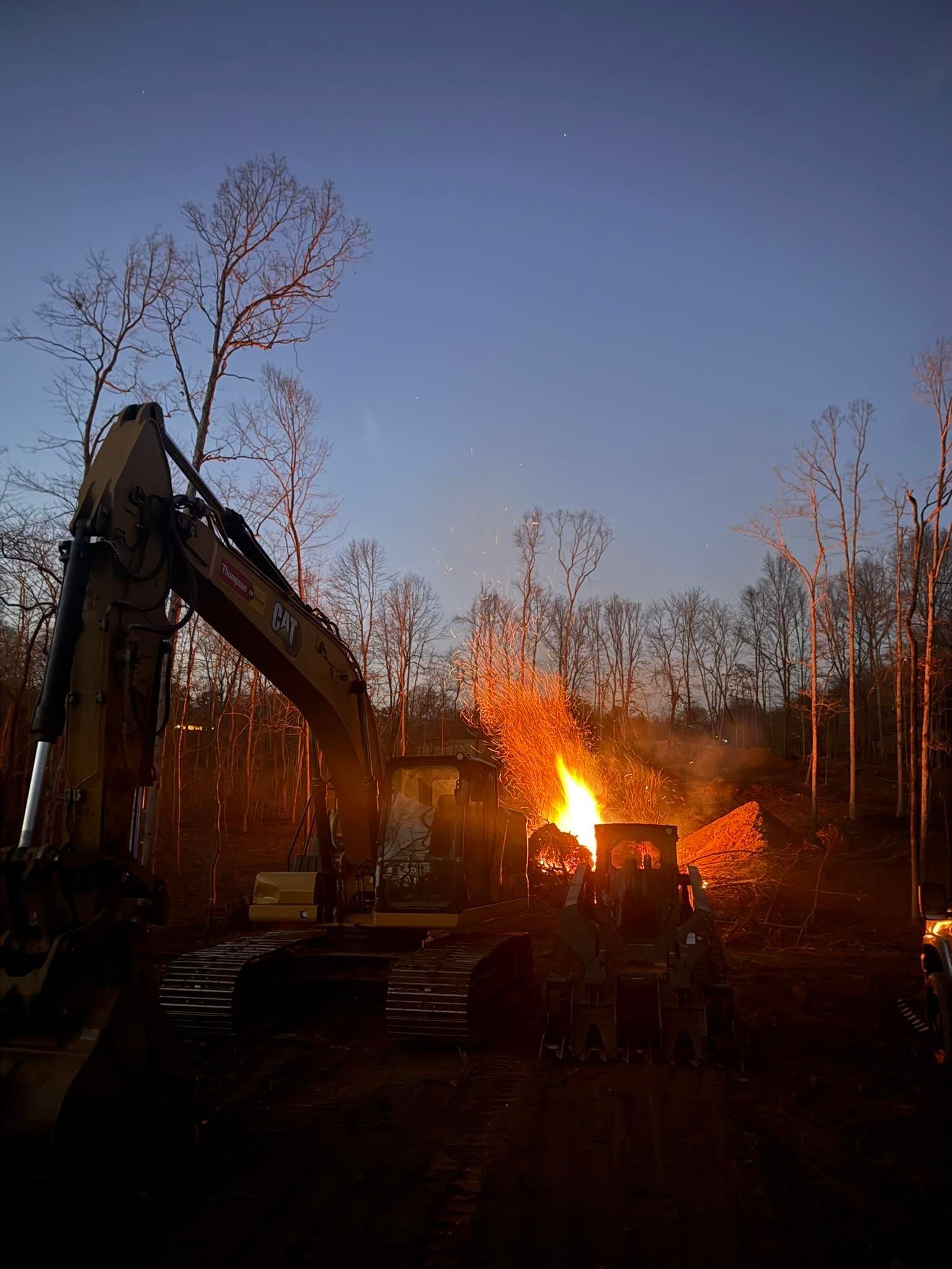 Excavator near a burning pile at dusk in a wooded area, with bright flames against the dark sky