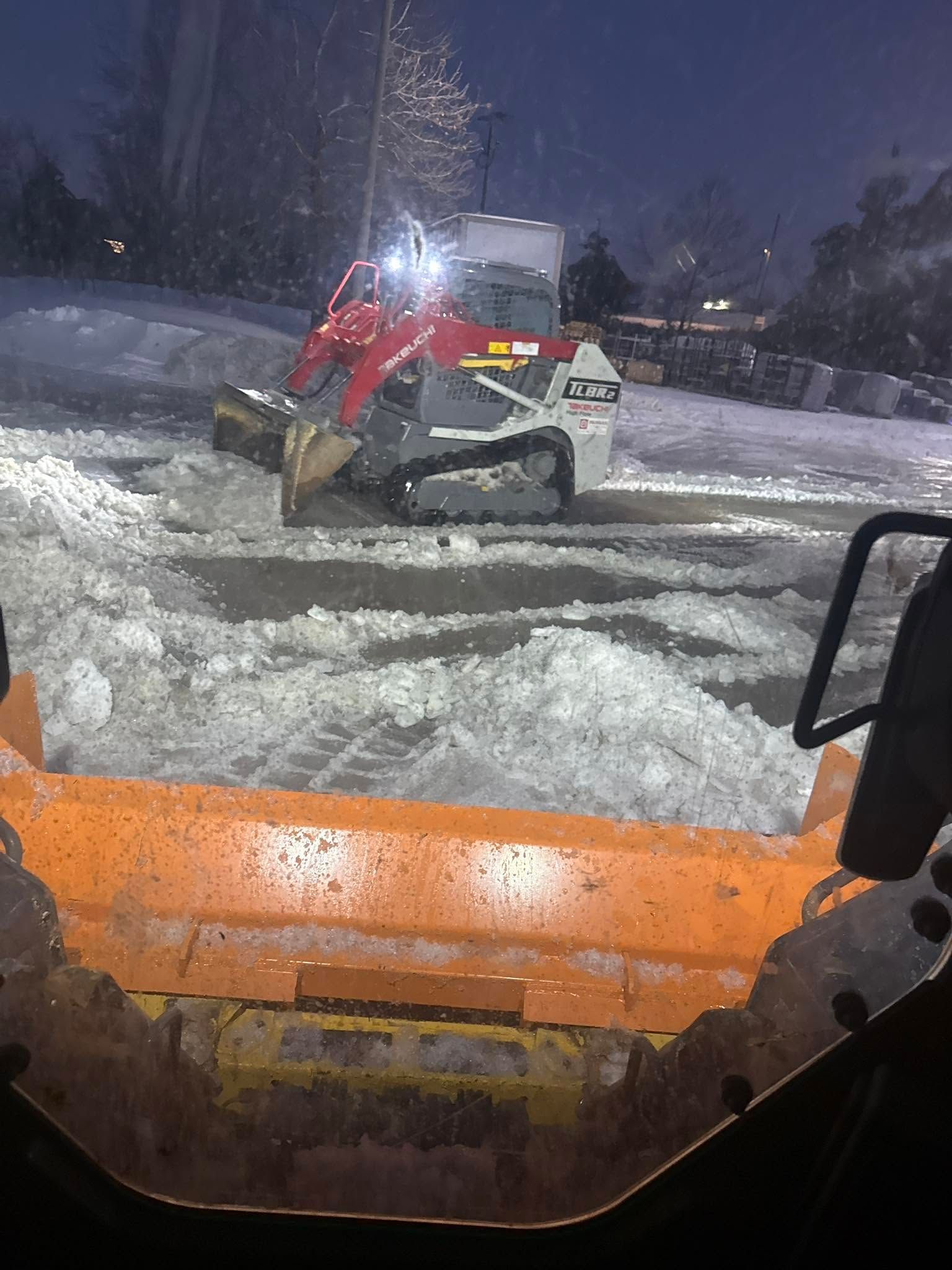 Snowplow clearing a snowy lot at night, seen through an orange vehicle windshield.