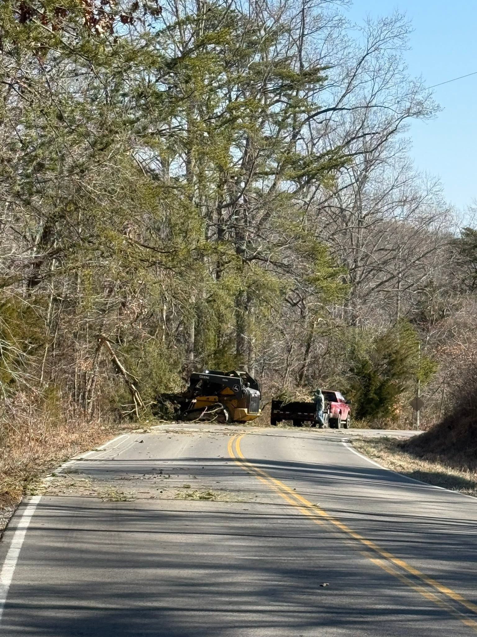 Blocked rural road with a yellow barrier and vehicles under trees in daylight