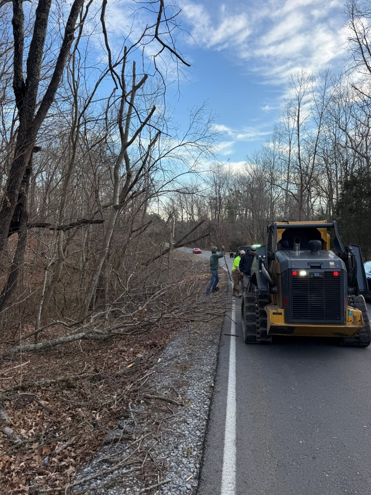 Workers and a backhoe clearing debris along a wooded roadside under a partly cloudy sky