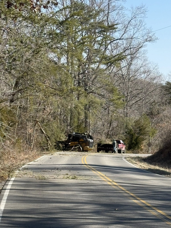 Rural road blocked by a fallen tree, with a work vehicle and a person clearing debris.
