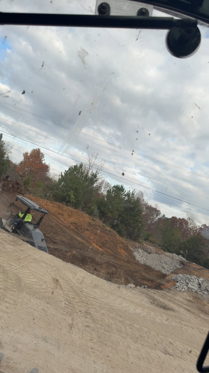 Construction site with excavator on a dirt pile under a cloudy sky, viewed through a vehicle window.