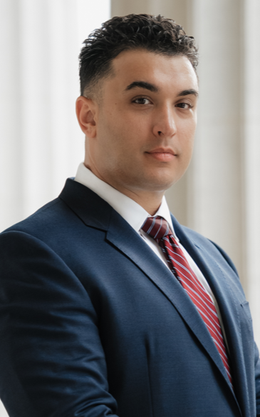 Man in a navy suit and red tie standing by a window with white curtains