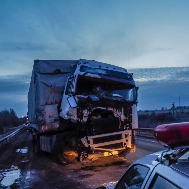 Damaged semi-truck on a road at dusk, with a police car in the foreground and smoke or steam rising nearby