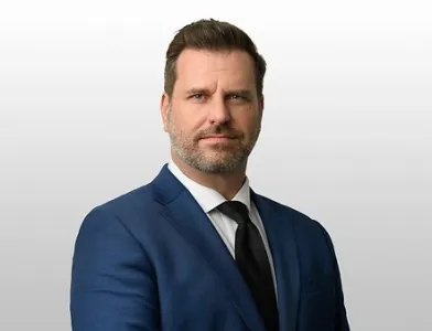 Man in a dark suit and tie, looking directly at the camera with a neutral expression.