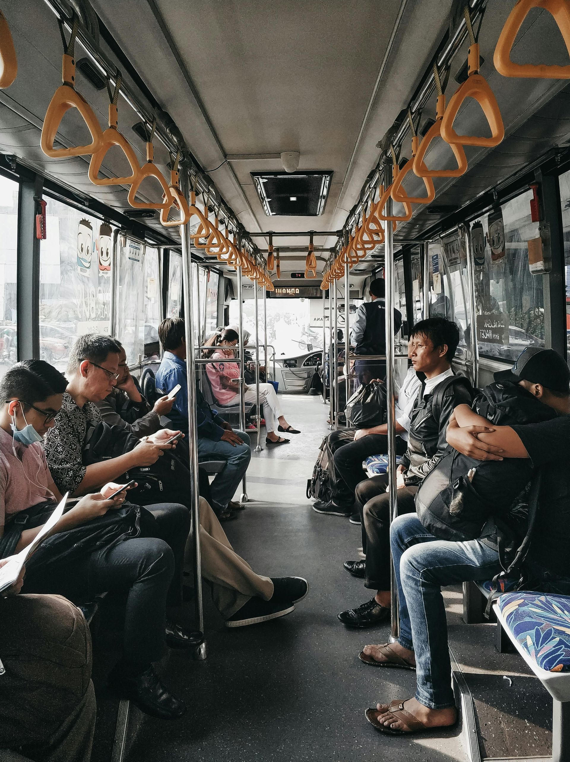 Passengers seated and standing in a crowded bus aisle, some looking at phones, with orange handrails overhead