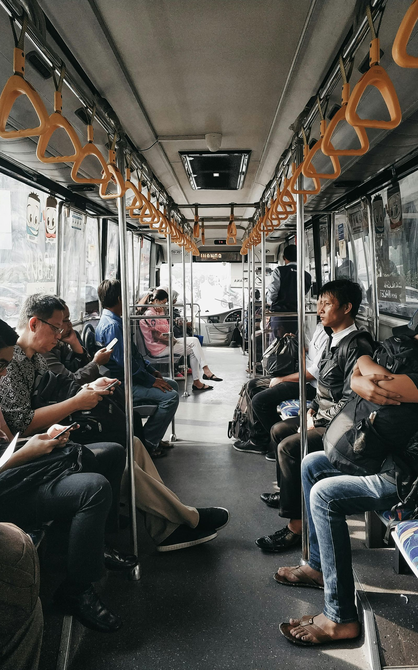 Interior of a bus, several people seated and standing. Overhead handrails are yellow. Sunlight streams in from windows.