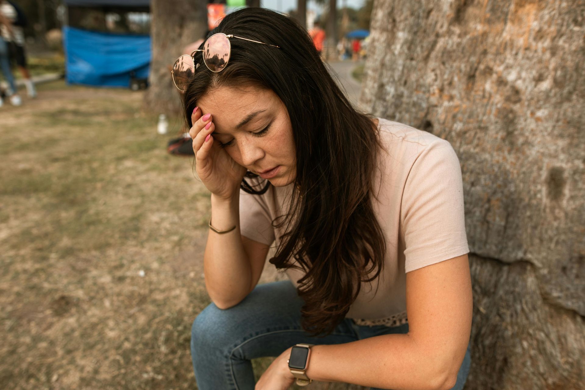 Woman sitting outdoors, holding her forehead and looking down, appearing stressed near a stone wall.