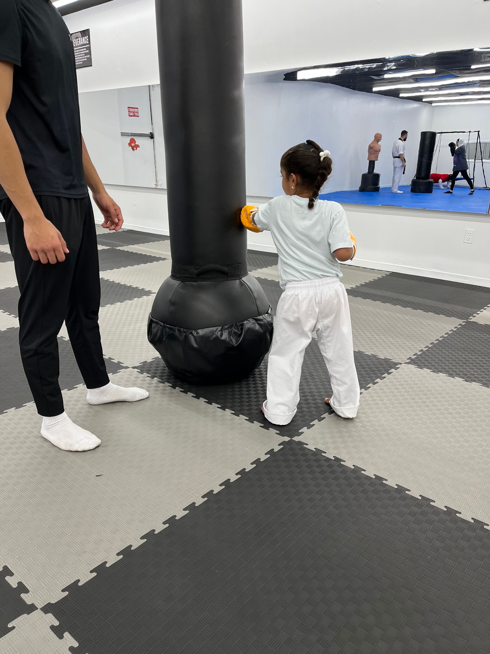 Child practicing martial arts in a gym beside a coach and a large black punching bag.