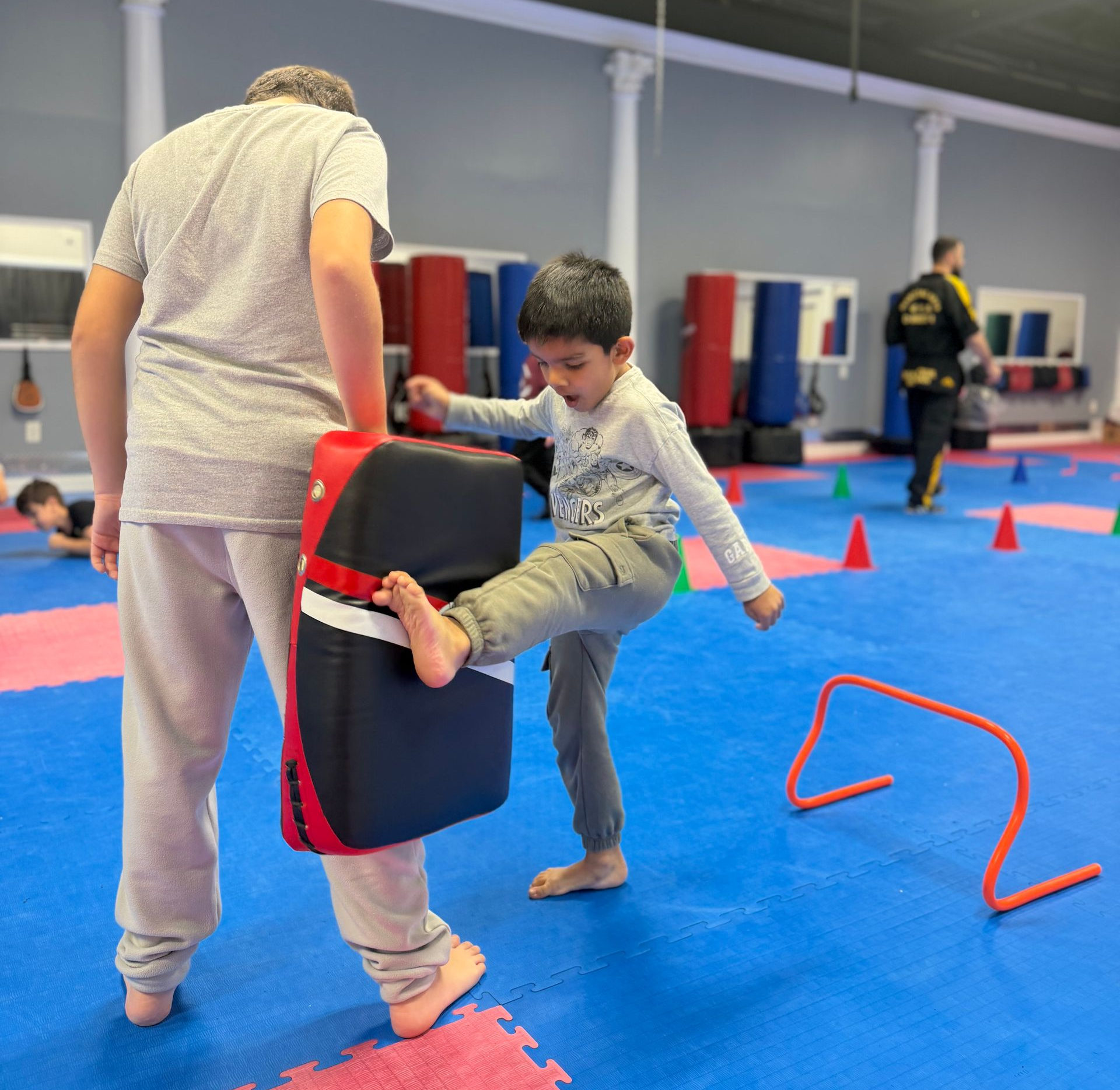 Child practicing martial arts kicks on a padded target in a blue training gym with cones and hurdles.