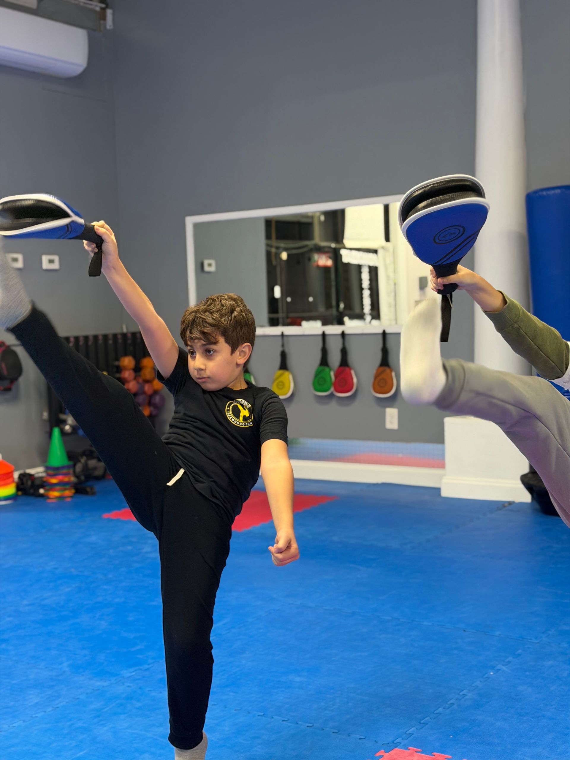 Child doing a high kick with a focus pad in a martial arts studio, another pad holder nearby