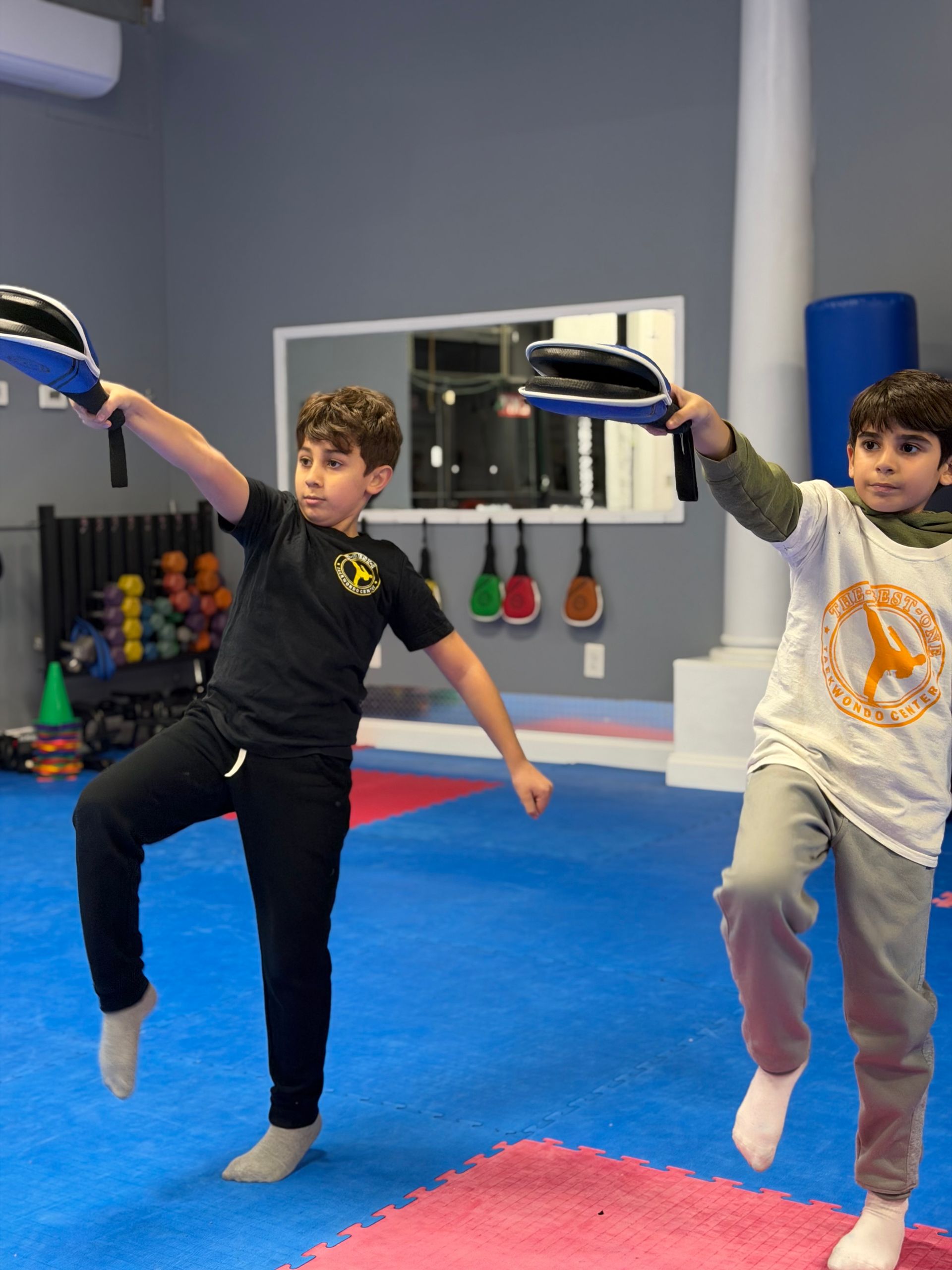 Two children doing balance drills on mats in a martial arts studio, each holding a pad up with one hand