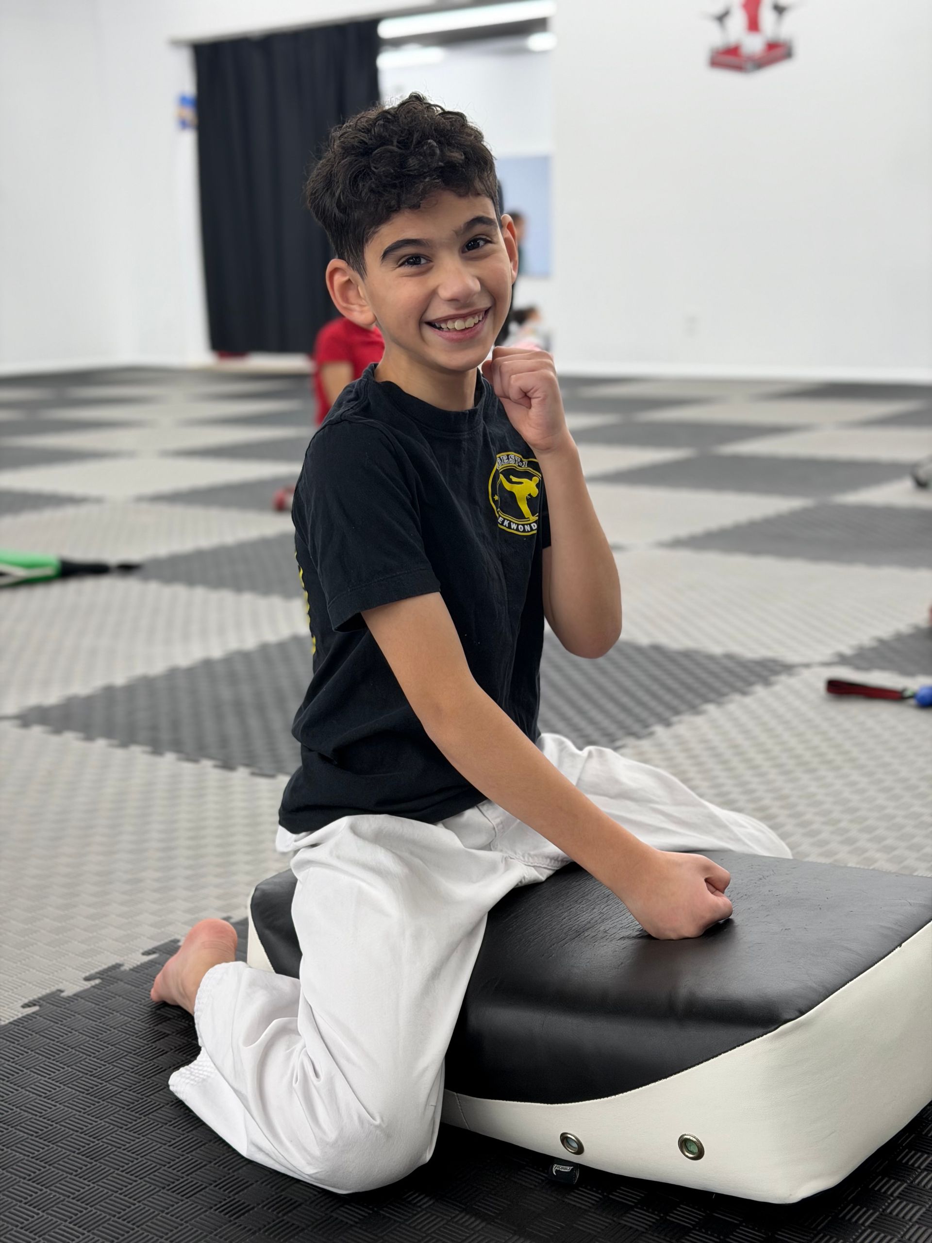 Smiling child kneels on a padded gym mat in a bright indoor room.