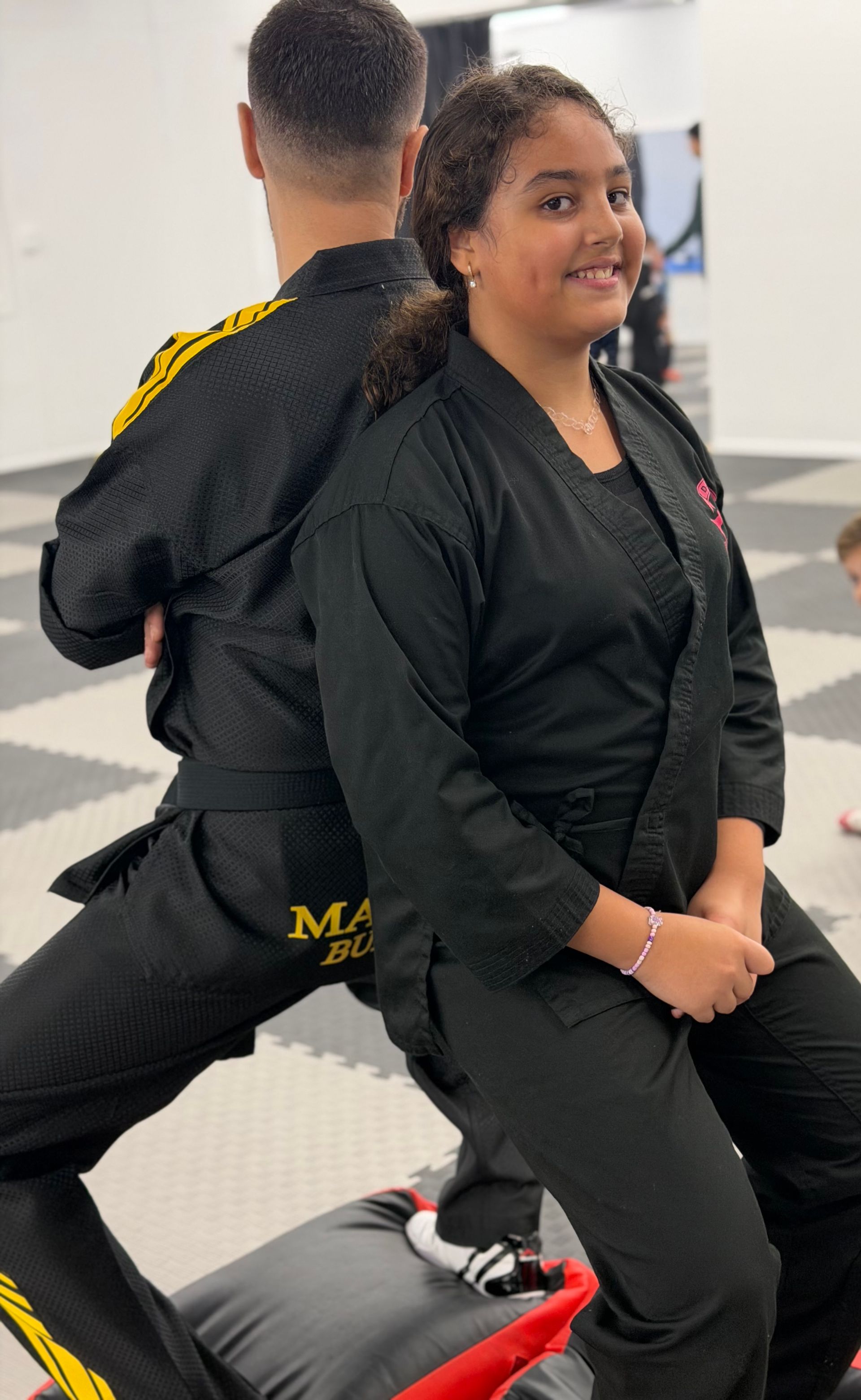 Two martial artists seated back-to-back on a mat, wearing black uniforms in a training studio