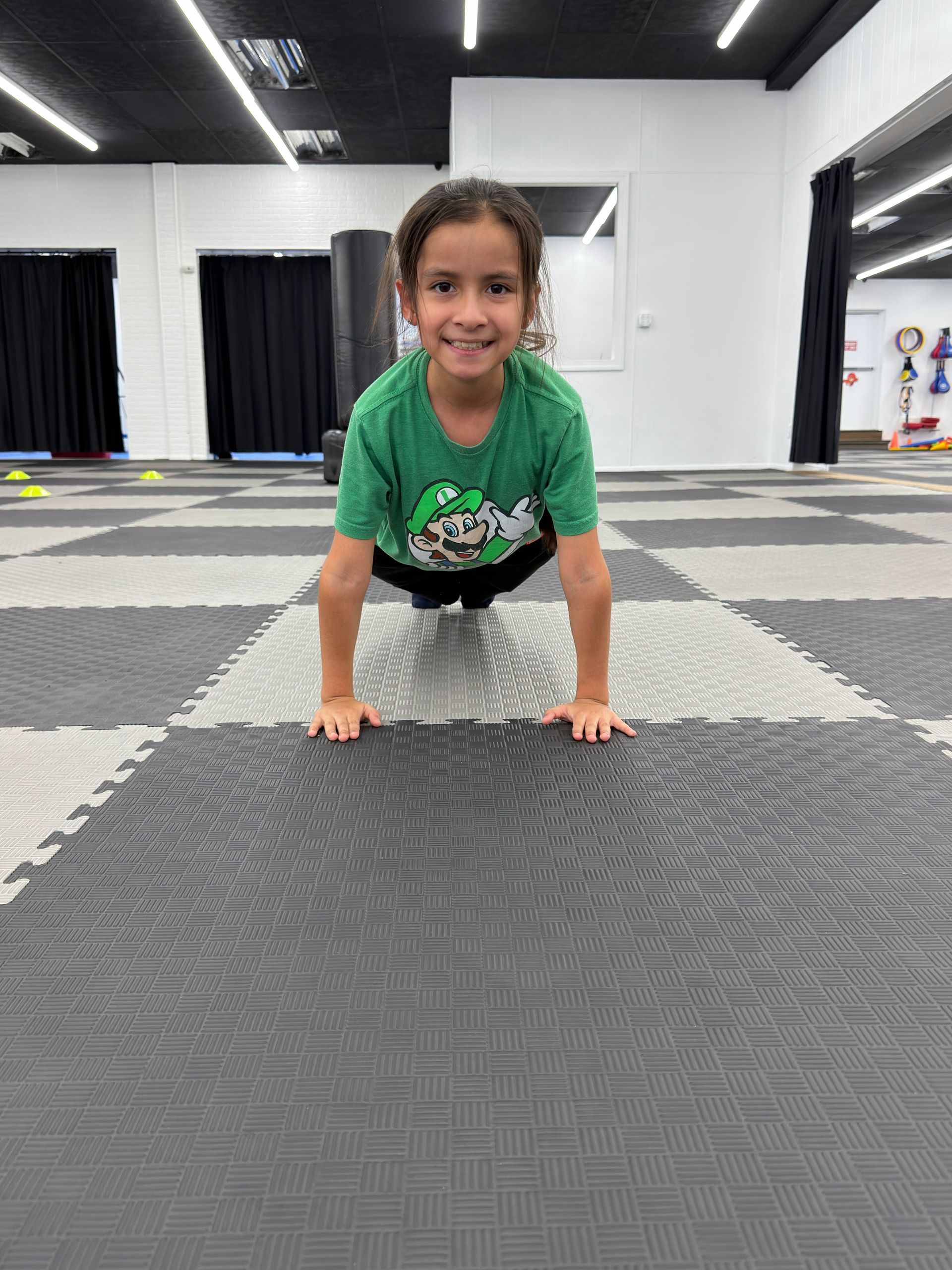 Smiling child in a green shirt doing a push-up on a gray gym mat in an indoor studio