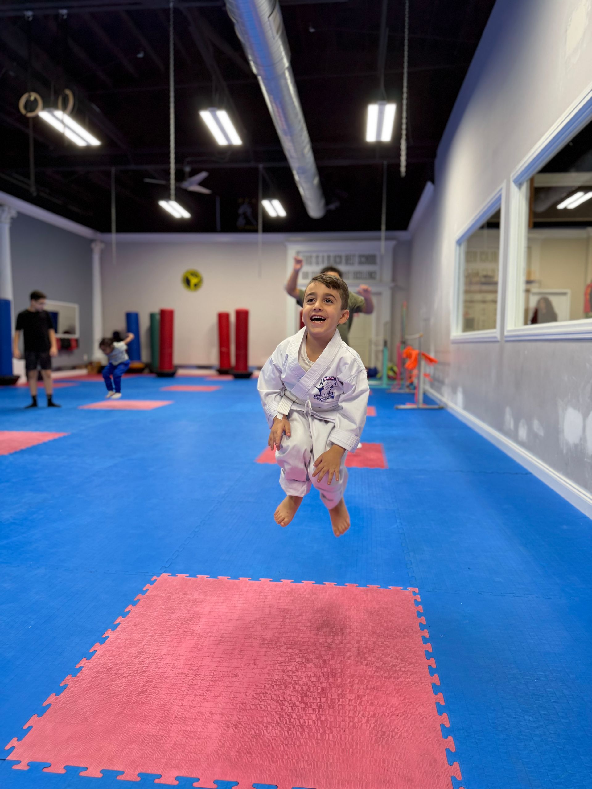 Child in a white gi jumping on blue gym mats in an indoor martial arts studio