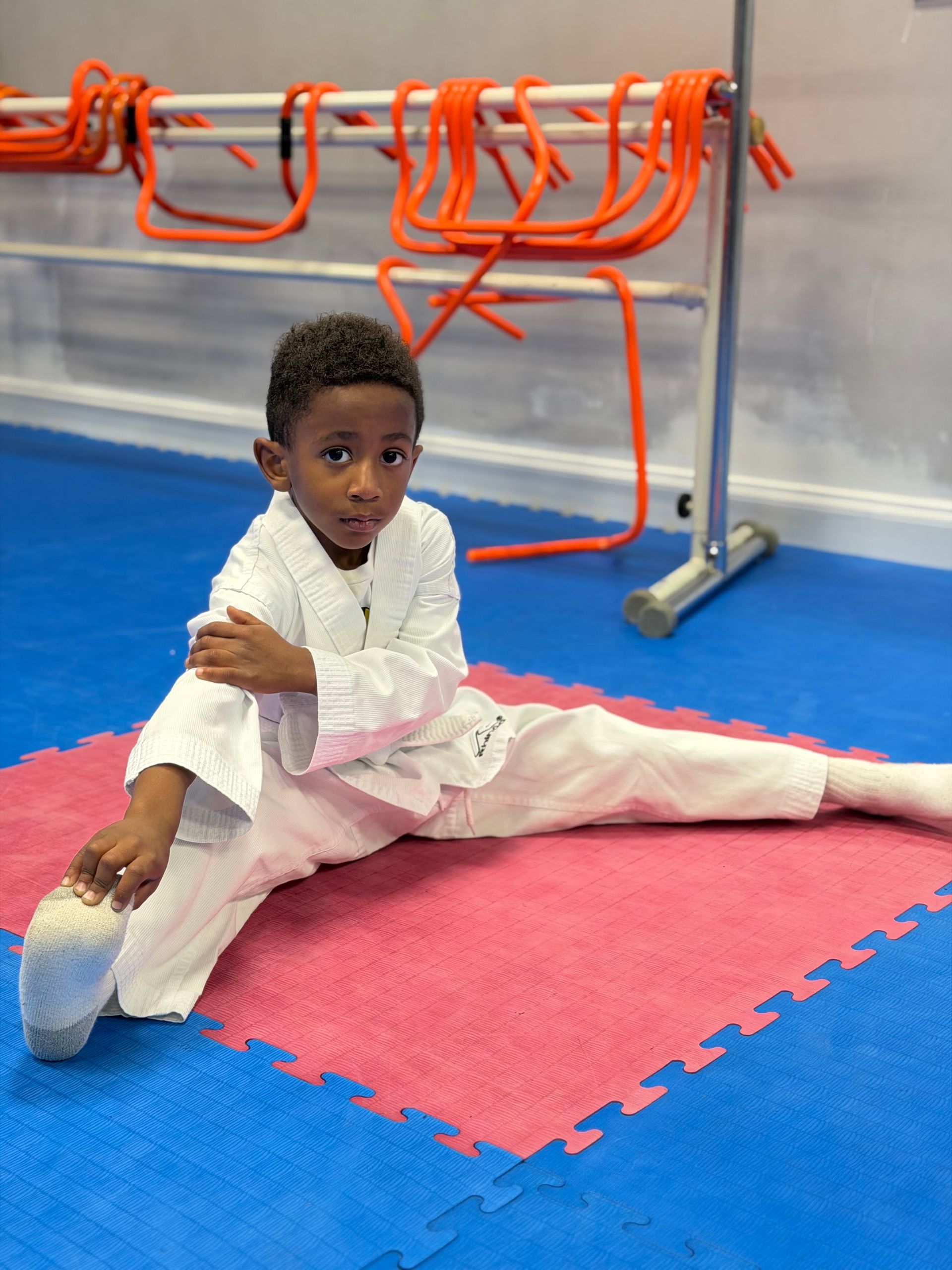 Child in a white outfit stretching on blue and pink mats in a gym, with orange hurdles in the background.