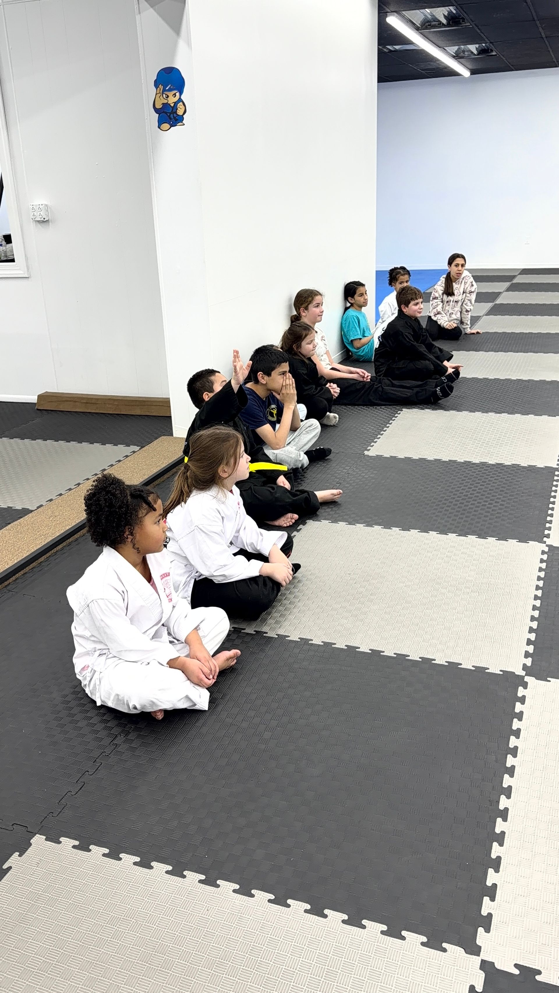People seated cross-legged on mats in a martial arts studio, lined up along a white wall.