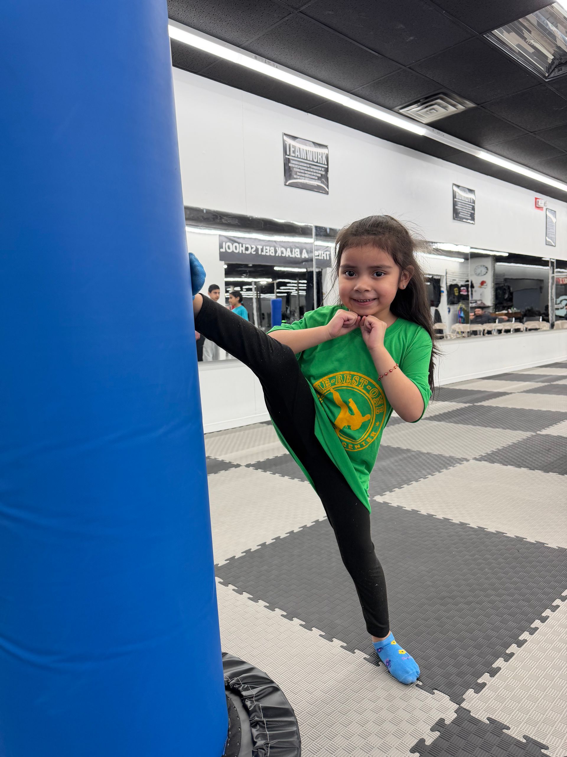 Child practicing a high kick beside a blue punching bag in a martial arts studio.
