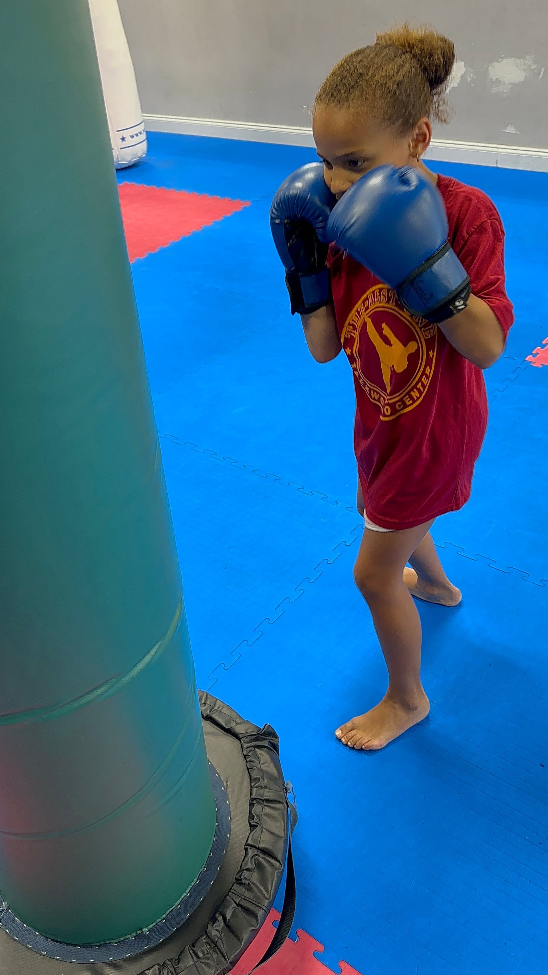 Child in blue boxing gloves and red shirt stands on a blue gym mat near a padded pillar.
