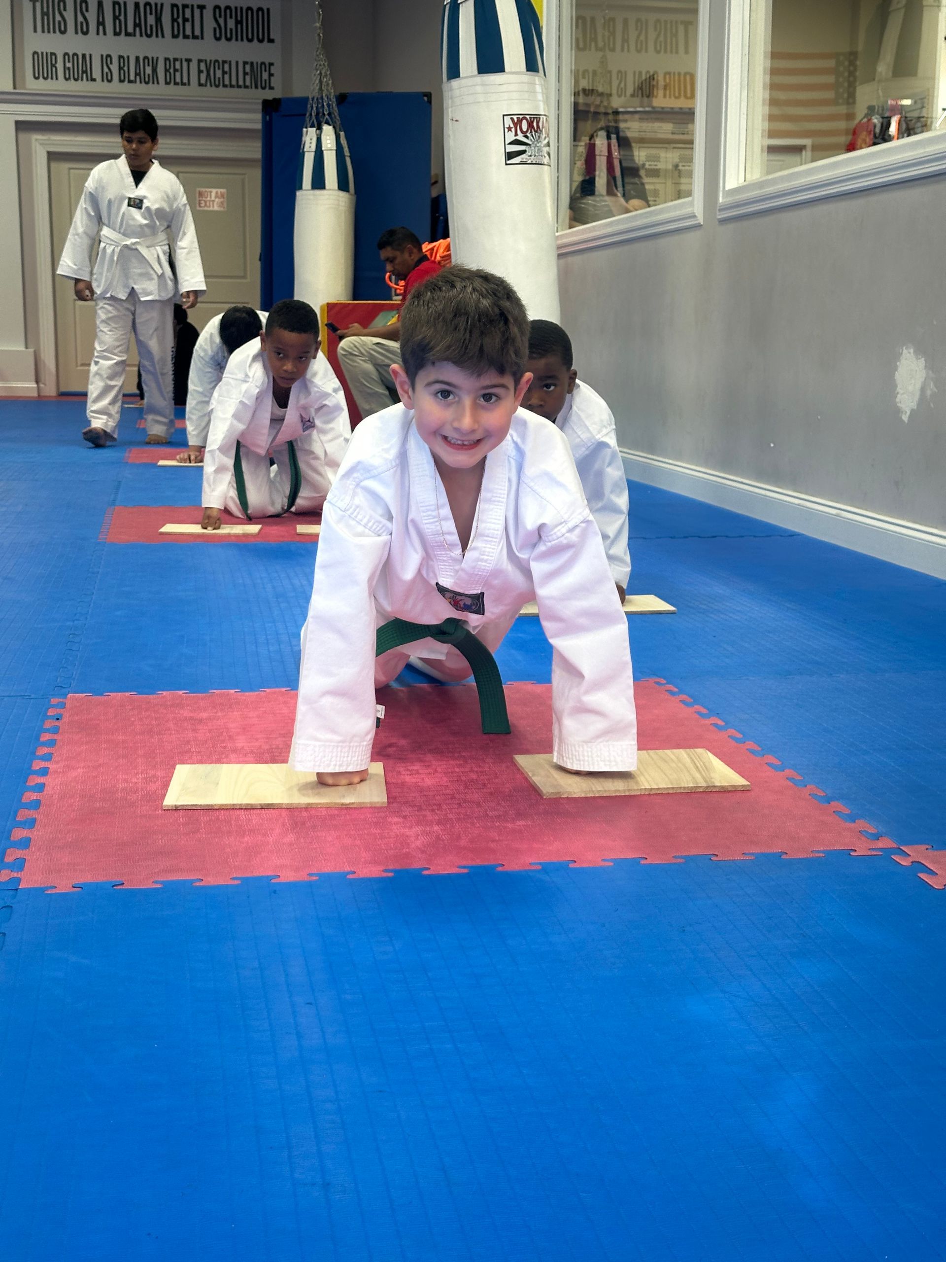 Children in white karate uniforms crawling on blue and red mats in a dojo, with an instructor in the background