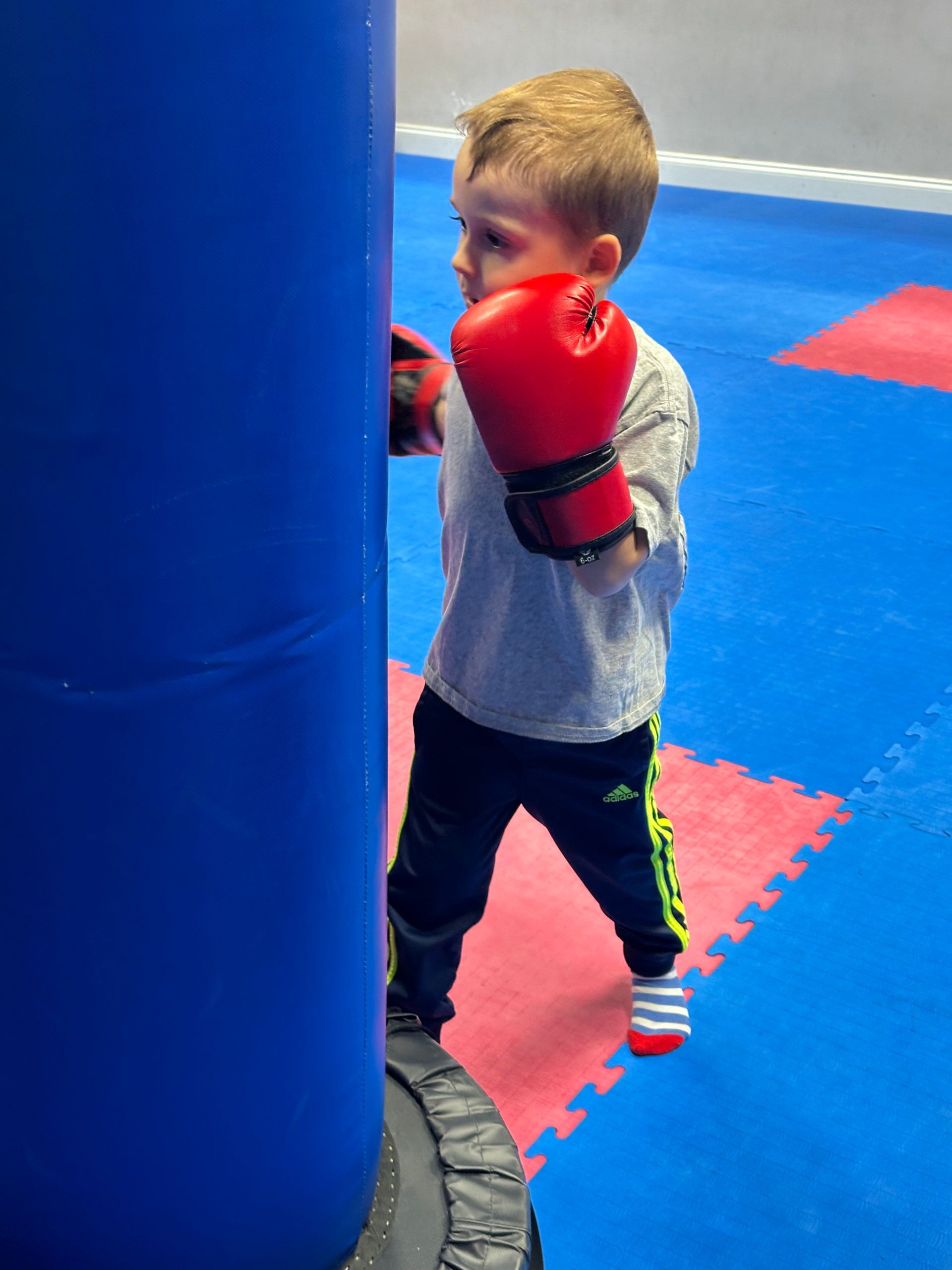 Child in red boxing gloves practicing punches beside a blue punching bag in a gym.