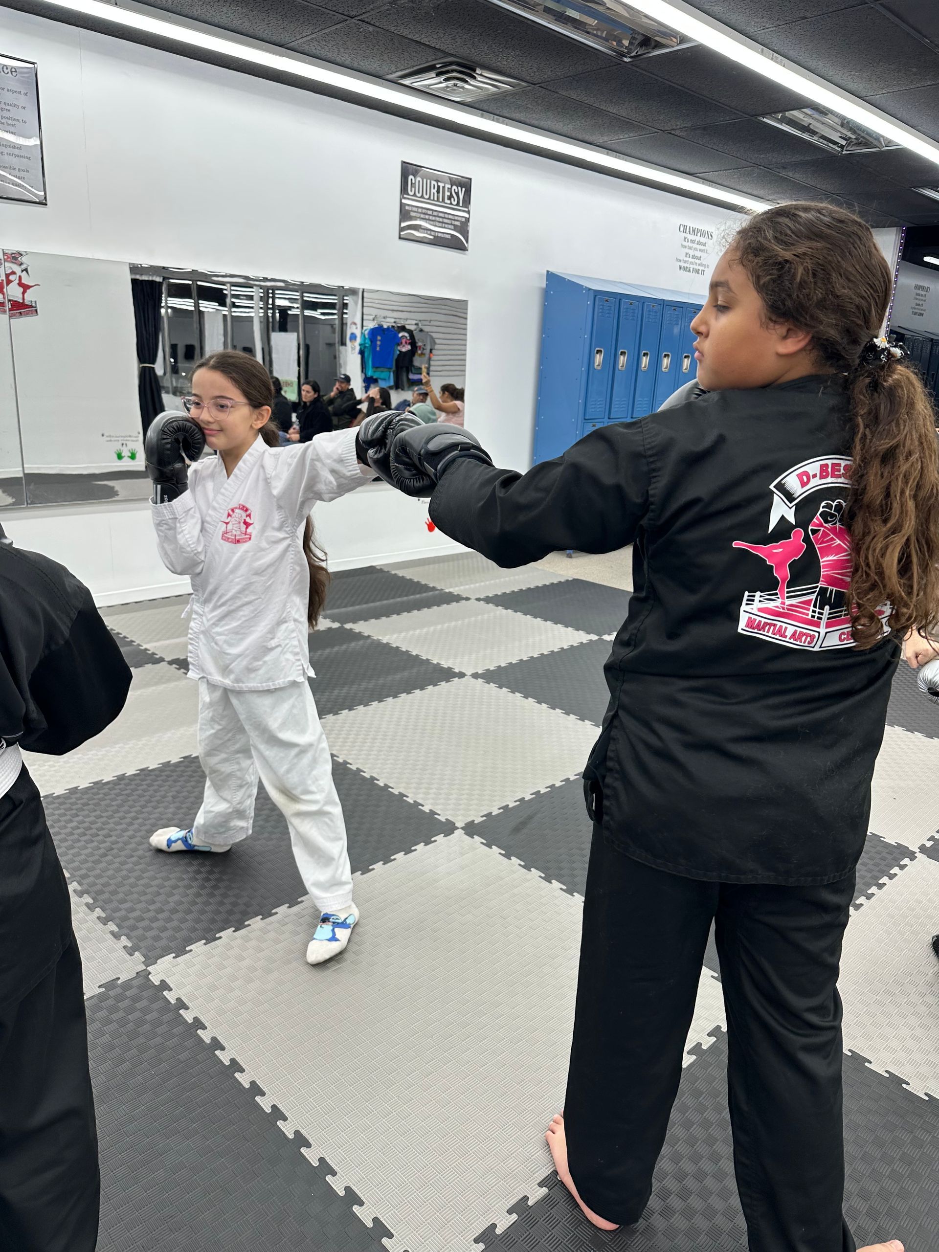 Two people practicing martial arts in a studio, one in white uniform, one in black, on a checkered mat