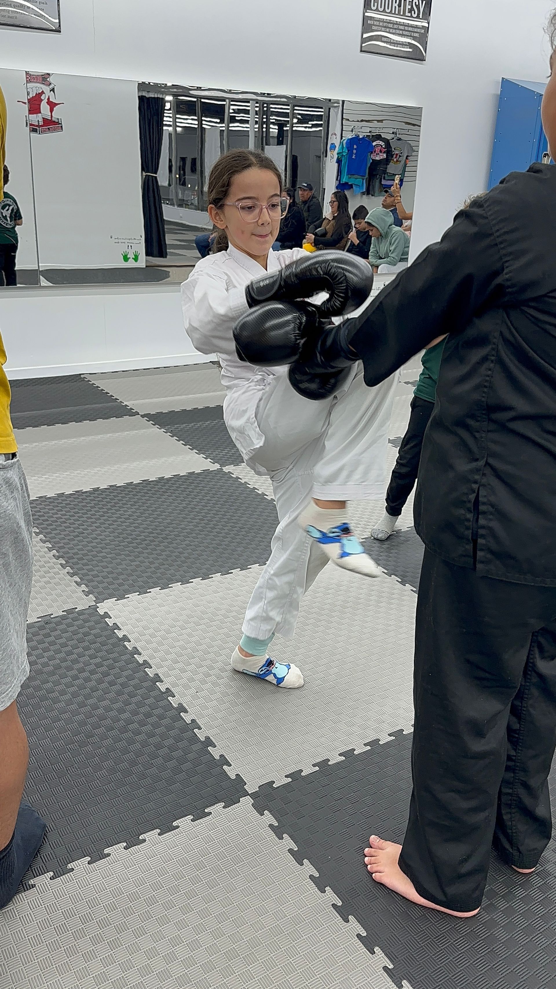 Child in a white martial arts uniform practicing with black-gloved instructor in a gym.