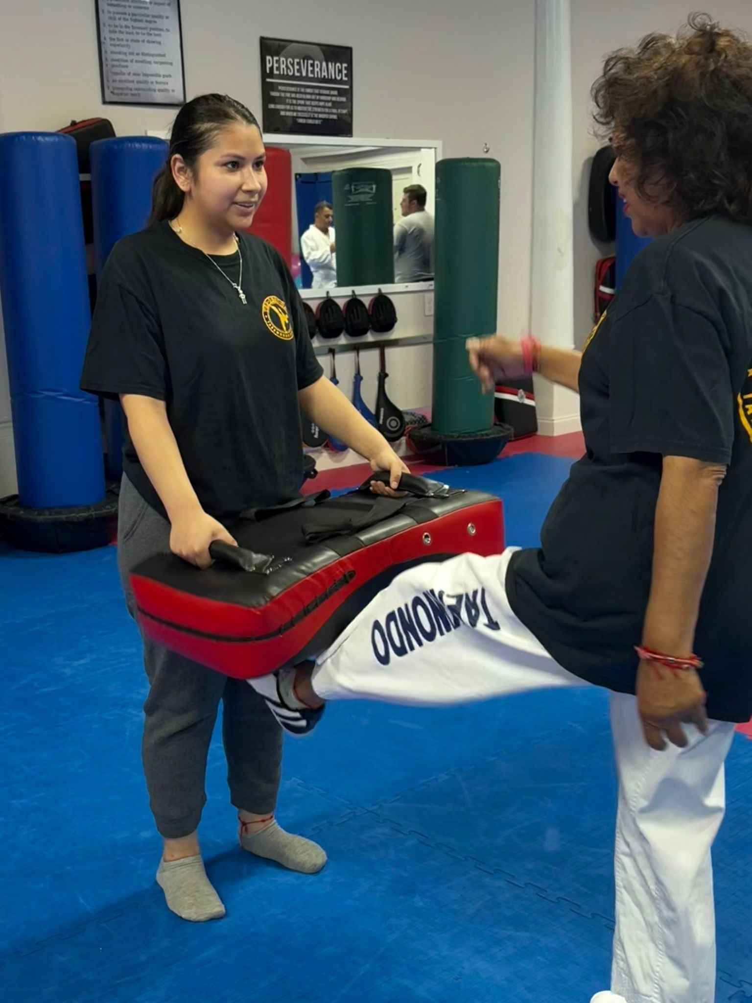 Two martial artists practice a kick shield drill in a gym, one holding a red-and-black pad.