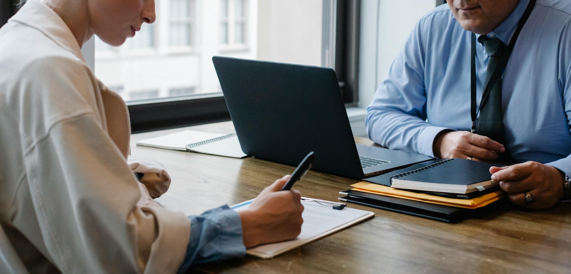 A man with a laptop and a woman signing a paper deposition are sitting at a table.