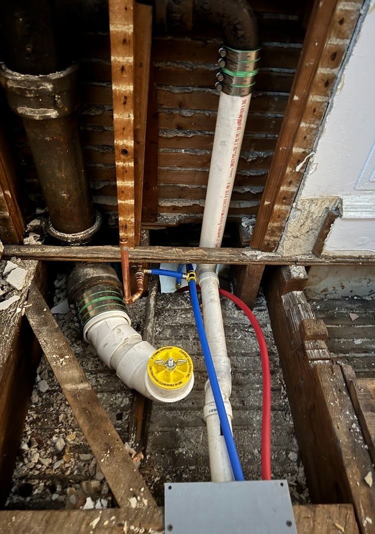 Plumbing pipes and fixtures exposed within a wooden ceiling: white, blue, red, and copper pipes, with a yellow cleanout cap.