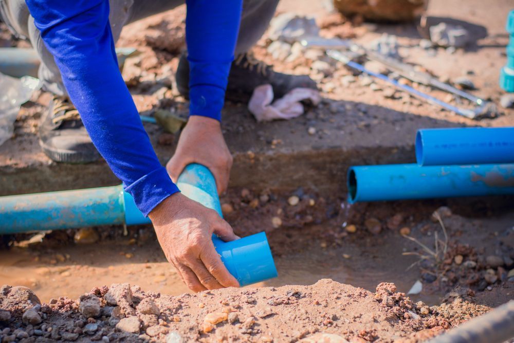 A person connecting blue PVC pipes in a dirt trench outdoors.