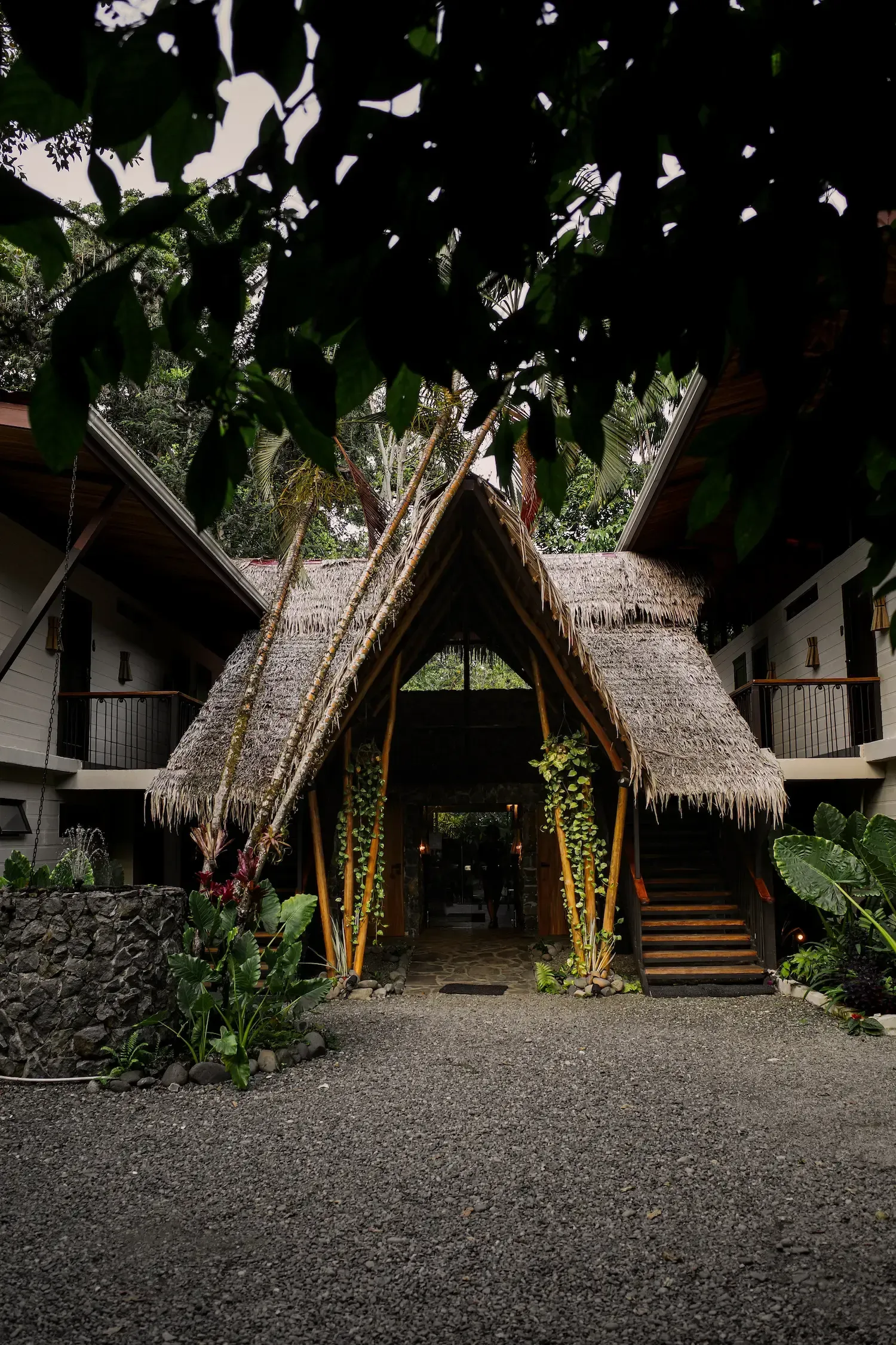 An open-air resort entrance with a thatched roof, surrounded by greenery and a gravel pathway.