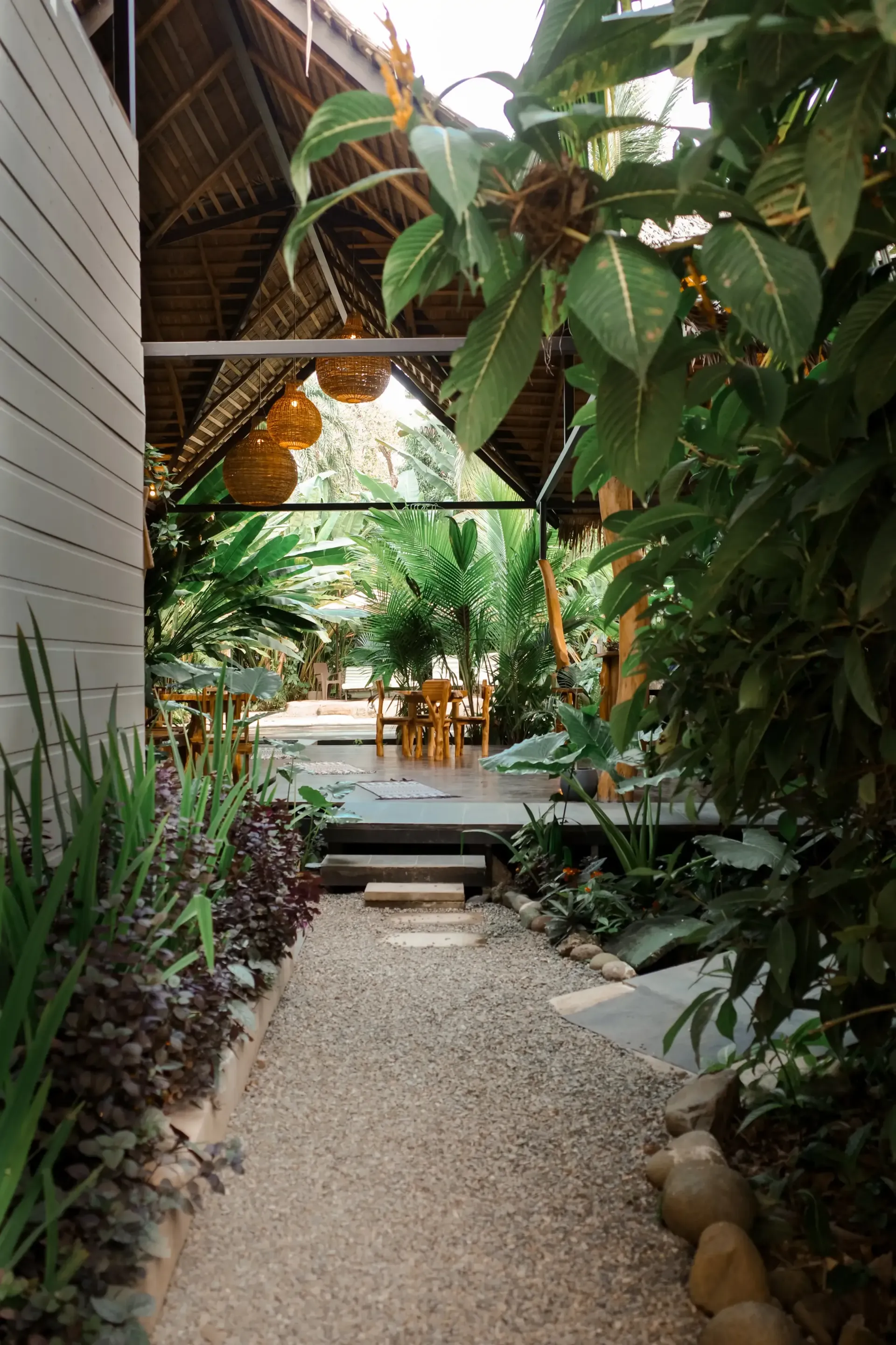 Gravel path leads to an open-air dining area under a thatched roof, surrounded by lush greenery.