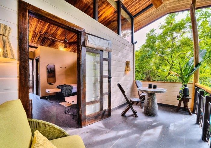 Balcony with a table and chairs, connected to a room with a bed, open to a lush green view.