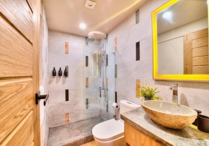 Bathroom with a wooden door, gray and stone walls, a shower, toilet, and sink with a yellow-framed mirror.