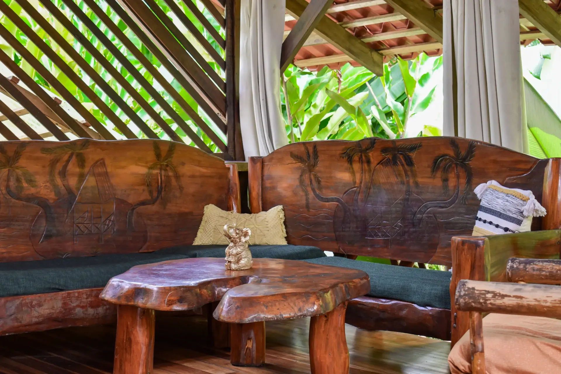 Wooden seating area with carved palm trees, a rustic table, and curtains, outdoors.