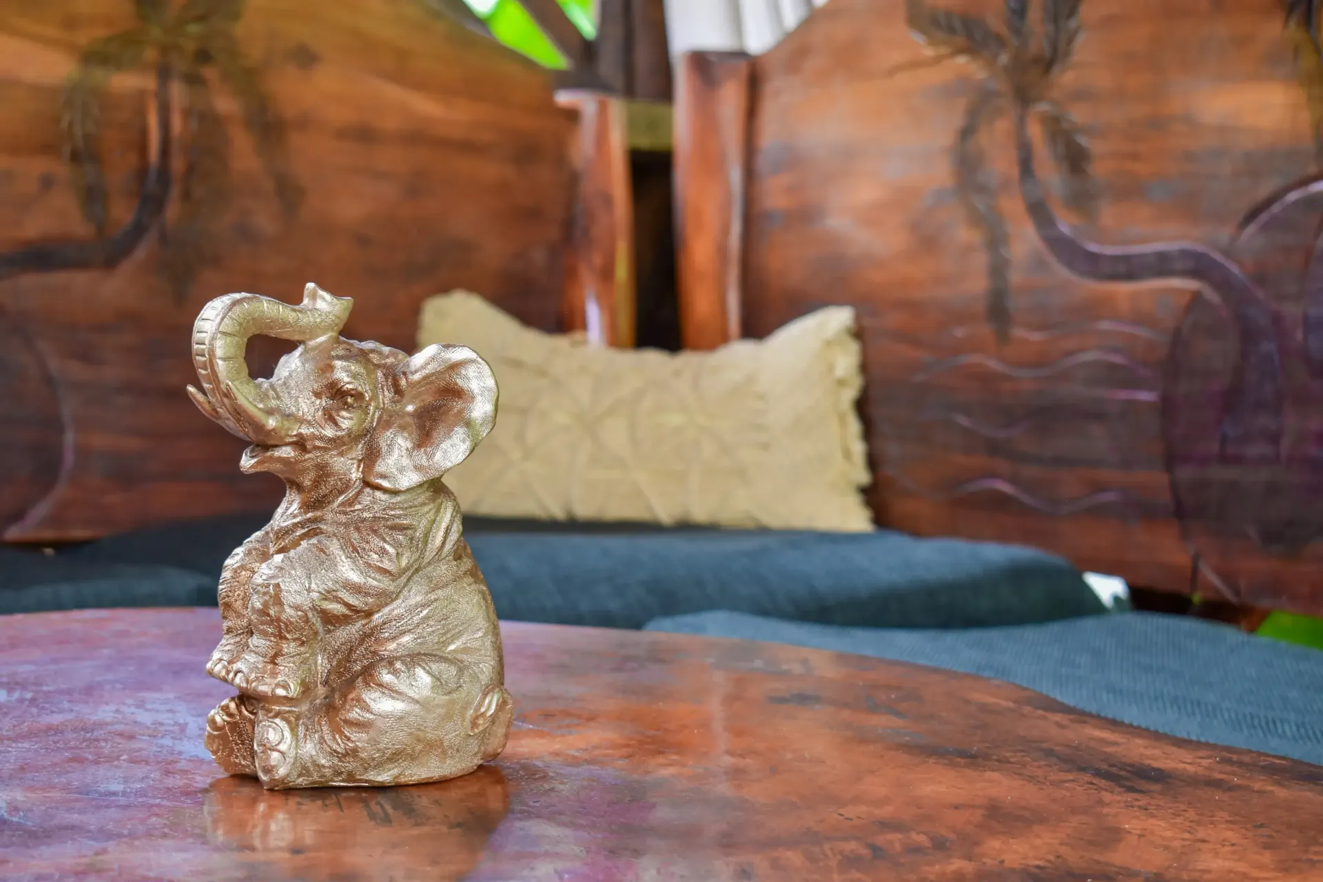 Gold elephant statue on a wooden table, with carved wooden furniture and a pillow in the background.