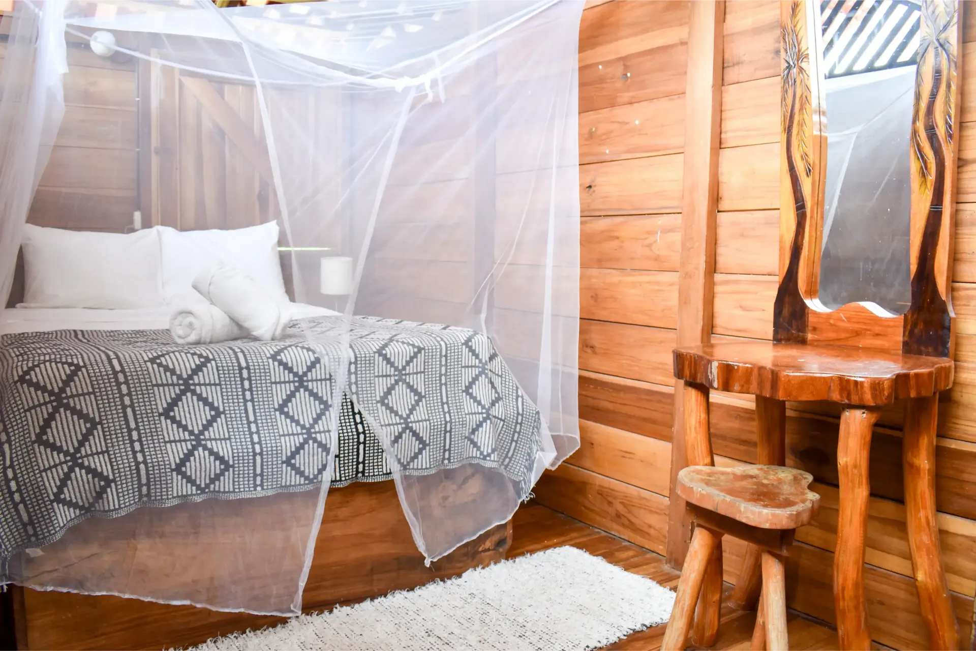 Bedroom with mosquito net over bed, wooden walls, vanity, and a small rug.