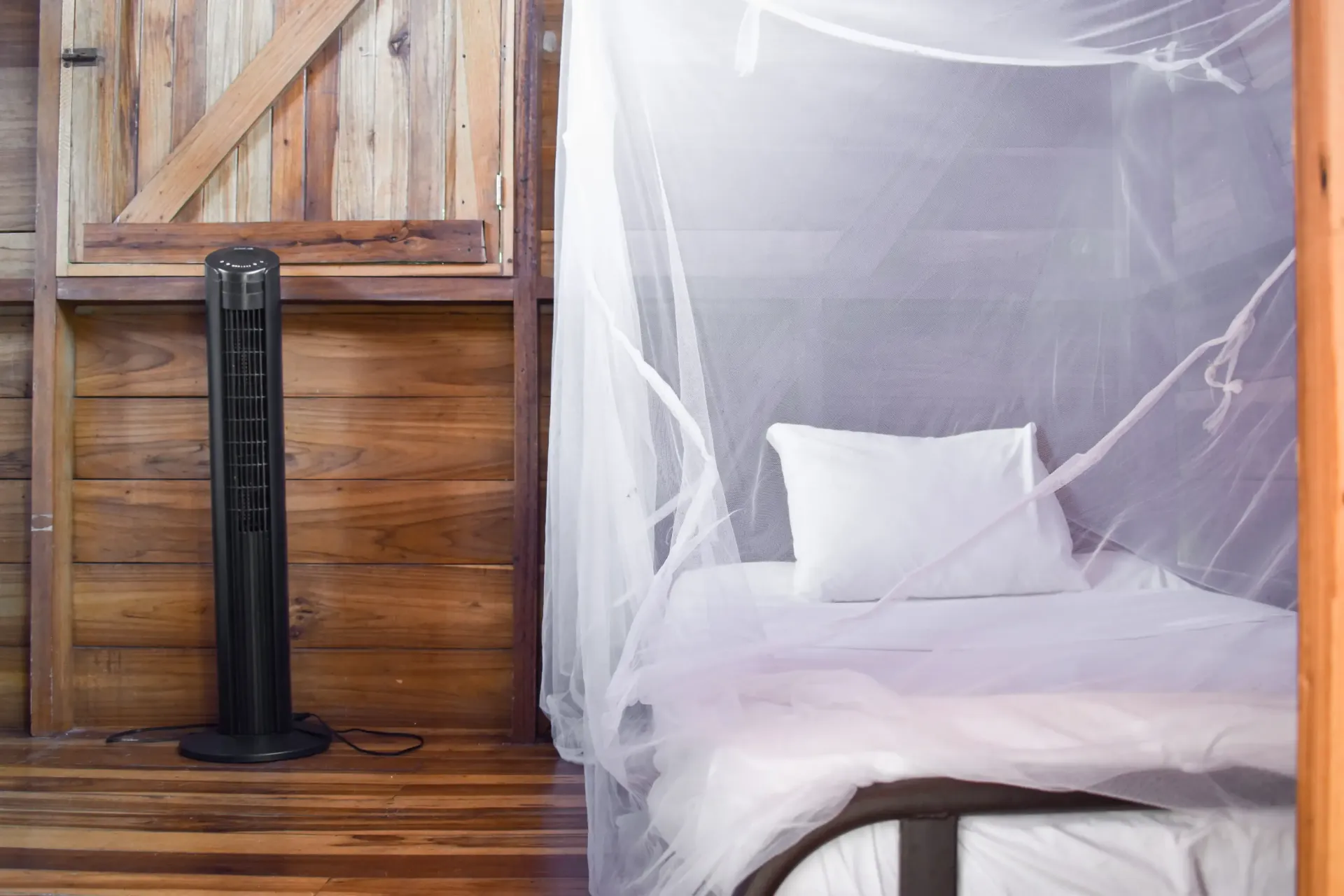 Black fan next to a bed with white mosquito netting, set in a wood-paneled room.