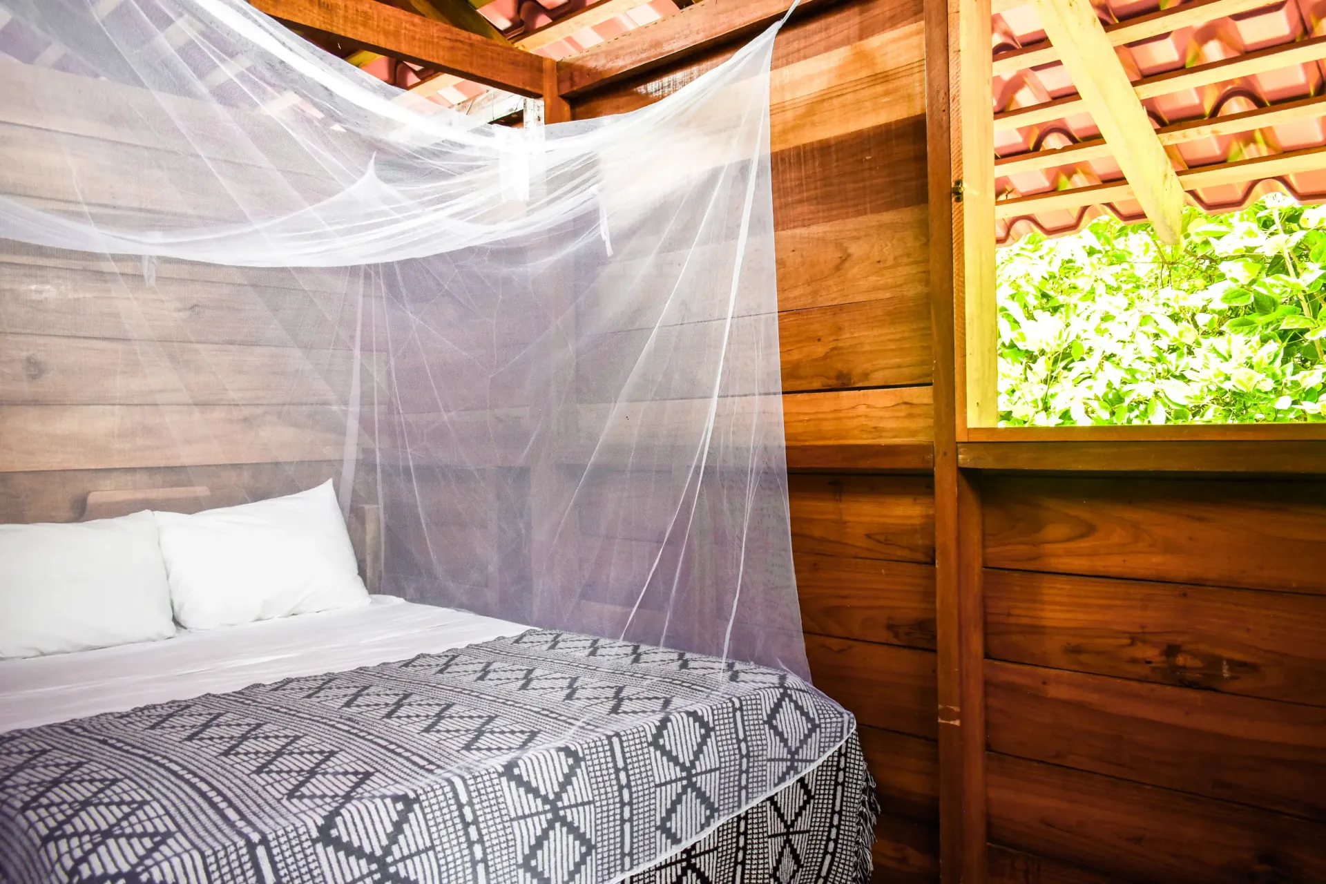 Wooden bedroom with mosquito net over bed; window reveals green foliage, red tile roof.