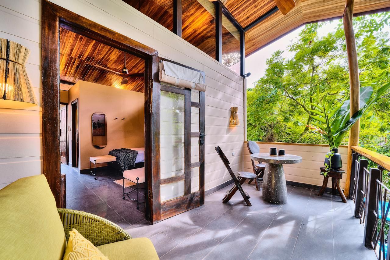 Balcony with table and chairs, leads to a bedroom. Wood and stone accents, surrounded by trees.