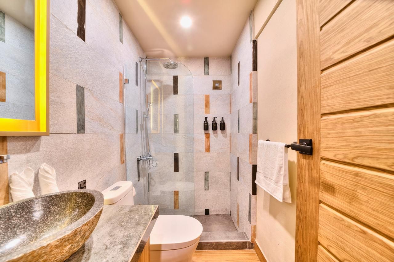 Bathroom with stone sink, toilet, and glass shower. Walls feature colorful rectangular tiles. Wooden door on right.
